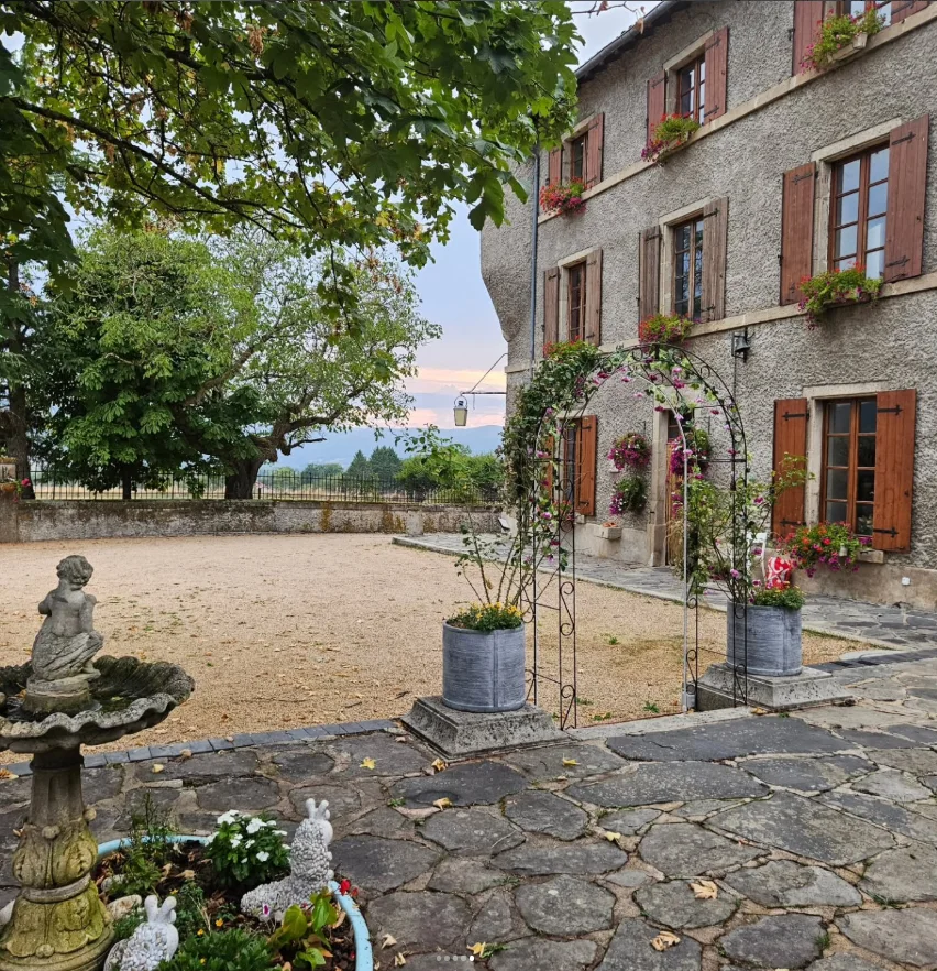 La cour du lieu d'accueil, à Beaulieu, en Haute-Loire : bâtisse en pierre aux volets bois, arche fleurie, fontaine et grand arbre au bord de la prairie.